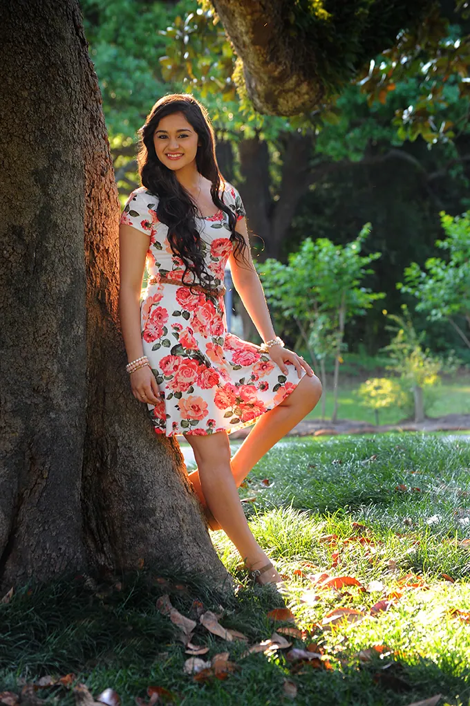 Senior girl in a floral dress standing in dappled sunlight beside a tree