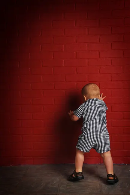 Toddler in a gingham romper standing against a red brick wall