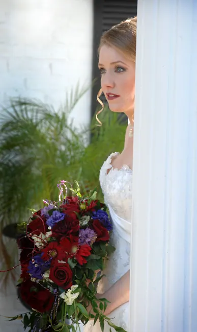 Bride gazing out a window holding a red and blue bouquet