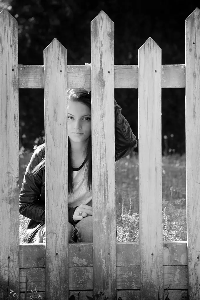 Black-and-white portrait of a senior girl peeking between white picket fence boards