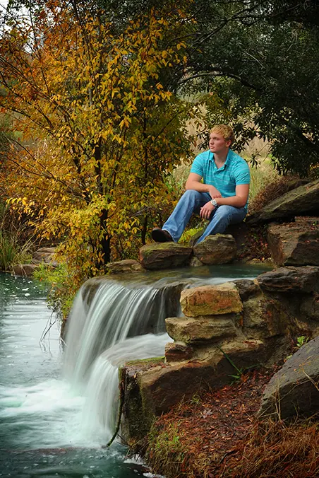 Senior boy seated on rocks above a small waterfall with autumn trees