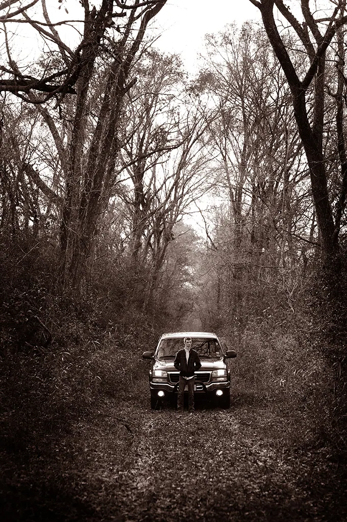Sepia portrait of a senior with his truck at the end of a wooded dirt road