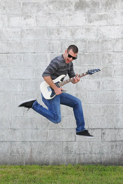 Senior guitarist in sunglasses leaping in midair against a white brick wall