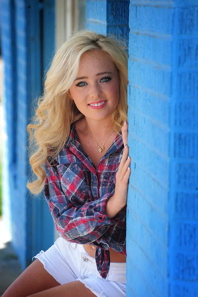 Blonde senior girl in a plaid shirt leaning against a bright blue wall