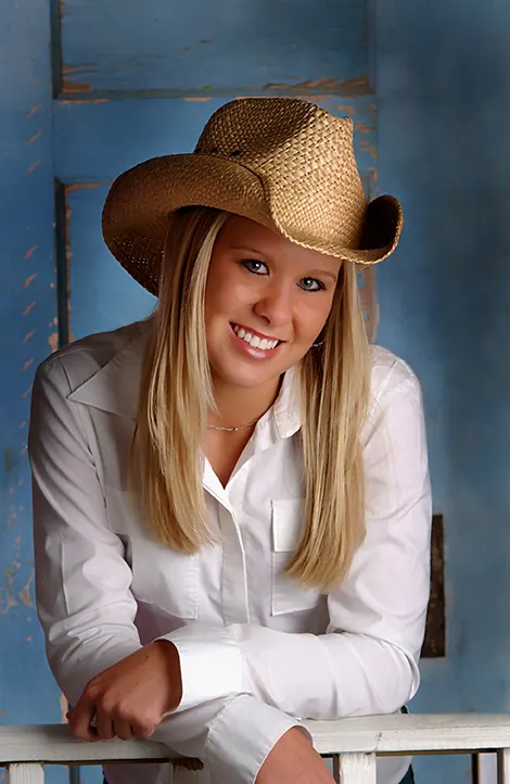 Smiling senior girl in a straw cowboy hat and white shirt leaning on a blue barn door
