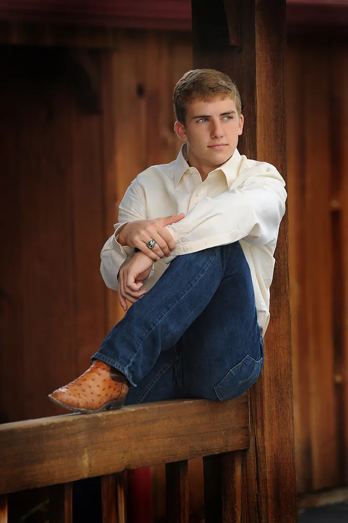 Senior boy in cowboy boots sitting on a rustic wooden fence rail