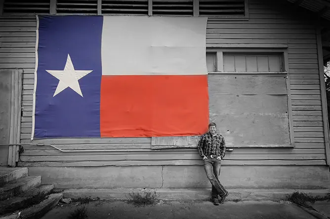 Senior in a cowboy hat sitting beneath a large Texas flag on a barn wall