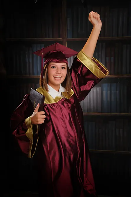 Graduate in a maroon cap and gown celebrating with a raised fist in a library