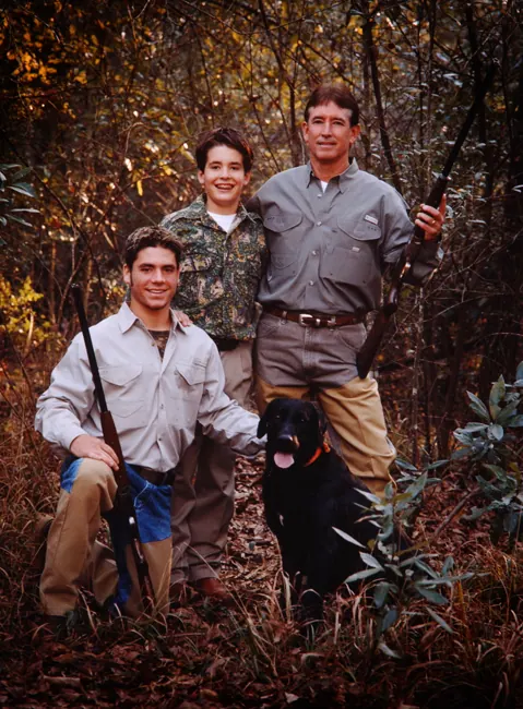 Father and sons with shotguns and a black Labrador in autumn brush