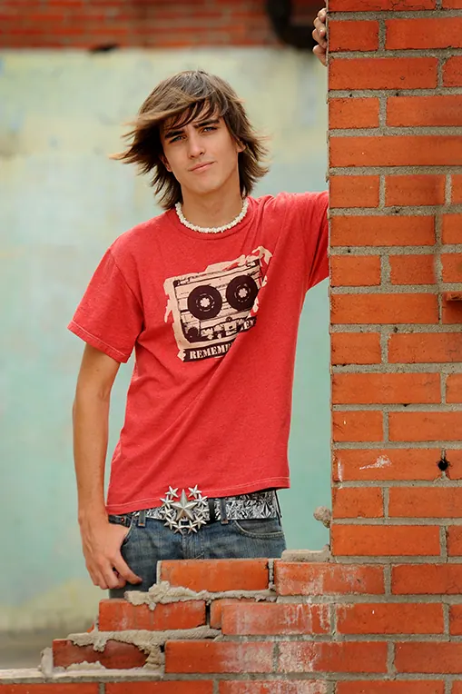 Teen boy in a red T-shirt and big belt buckle leaning on an old brick wall