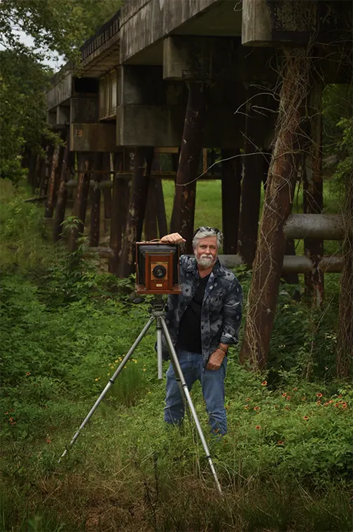 Jack Denlinger with a vintage large-format view camera on a tripod beneath a wooden trestle